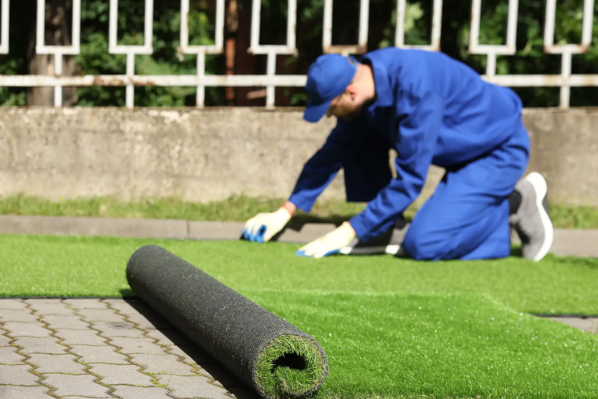 A person wearing blue work clothes and gloves is kneeling on the ground, installing artificial grass near a paved area, with a roll of synthetic turf in the foreground.