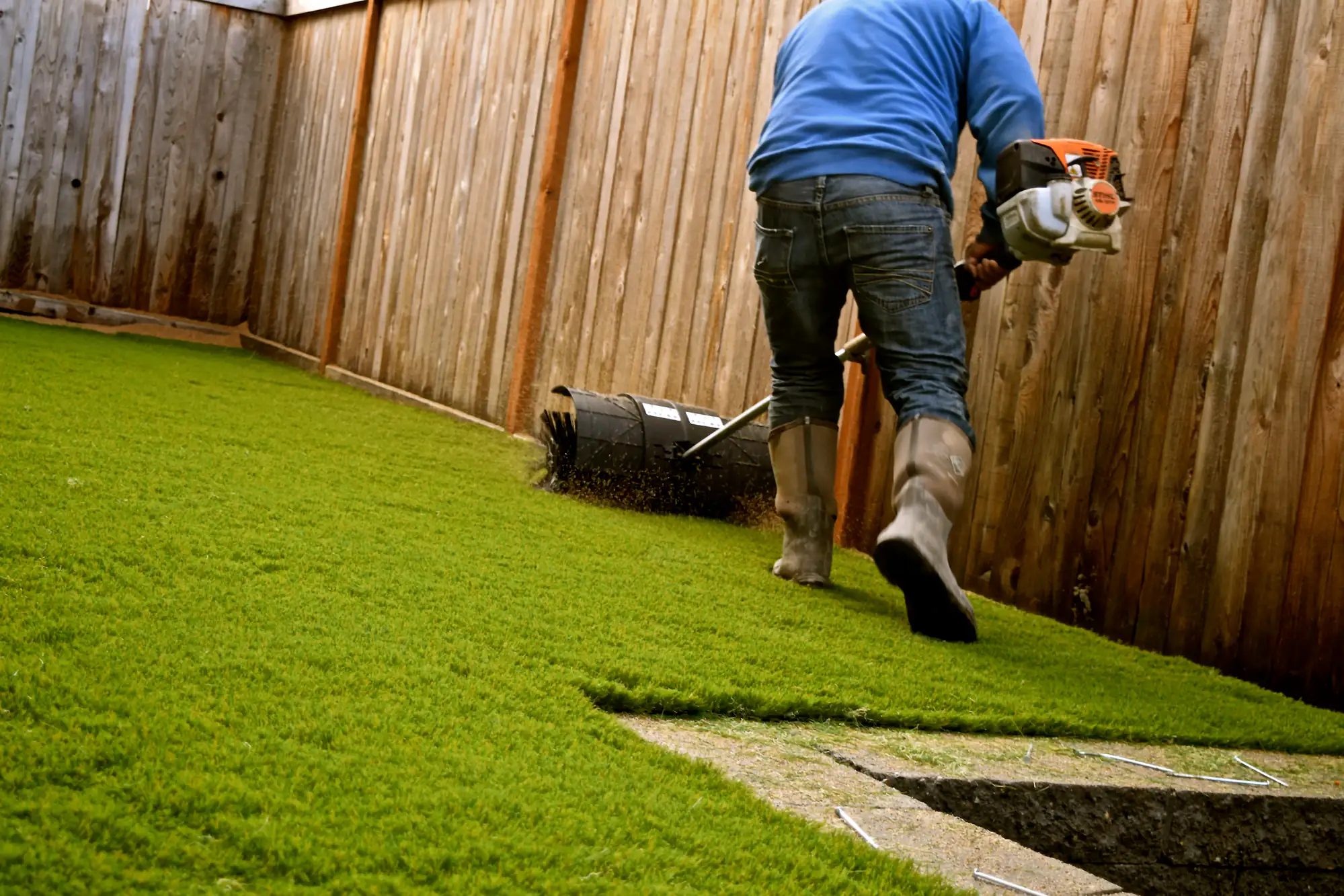 A person in jeans, boots, and a blue shirt is installing or grooming artificial grass in a fenced backyard using a power tool. A small section of grass is cut out, revealing the ground underneath.