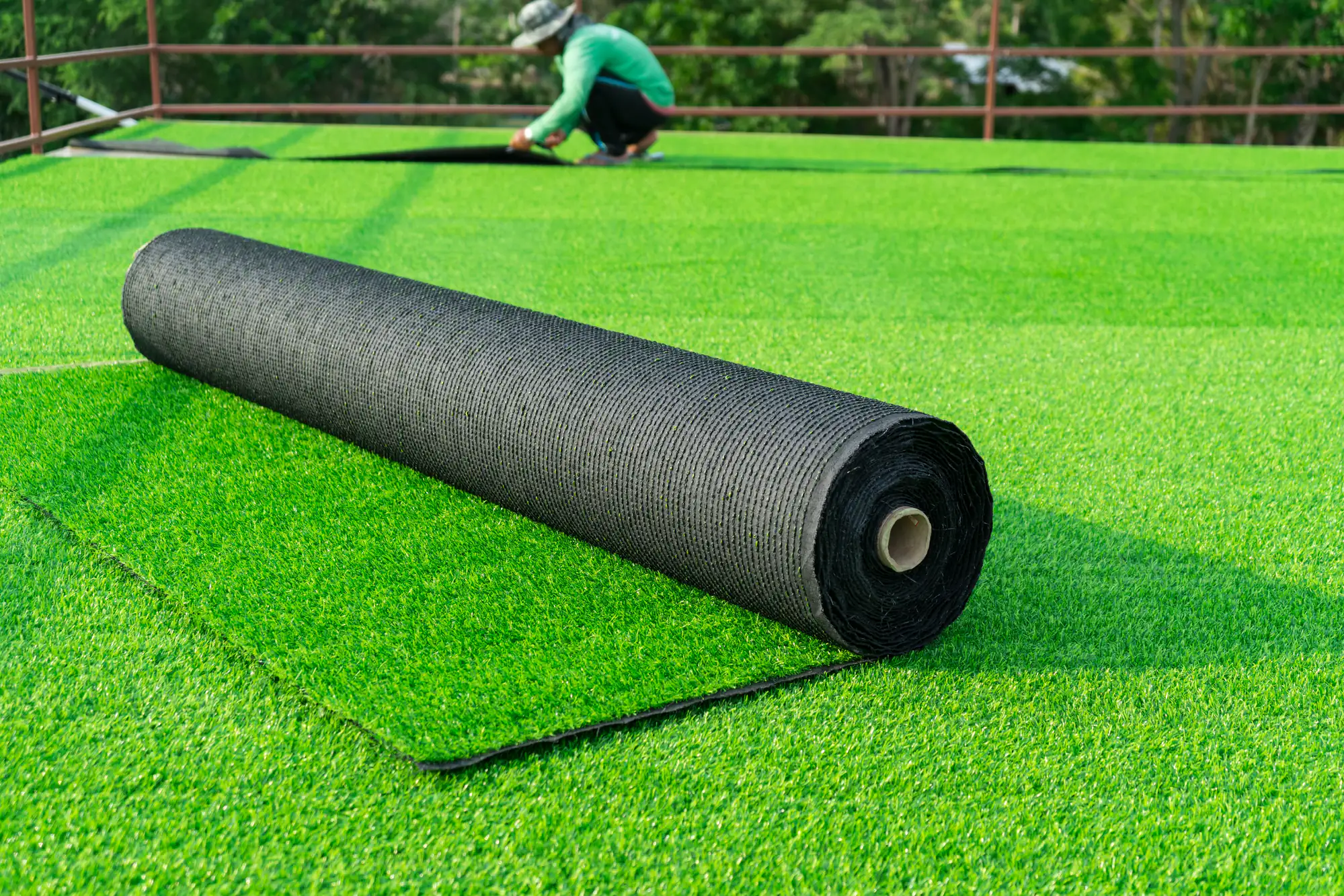 A large roll of artificial grass is partially unrolled on a field, with a person in the background fitting the turf. The scene is outdoors with green trees visible behind a fence.