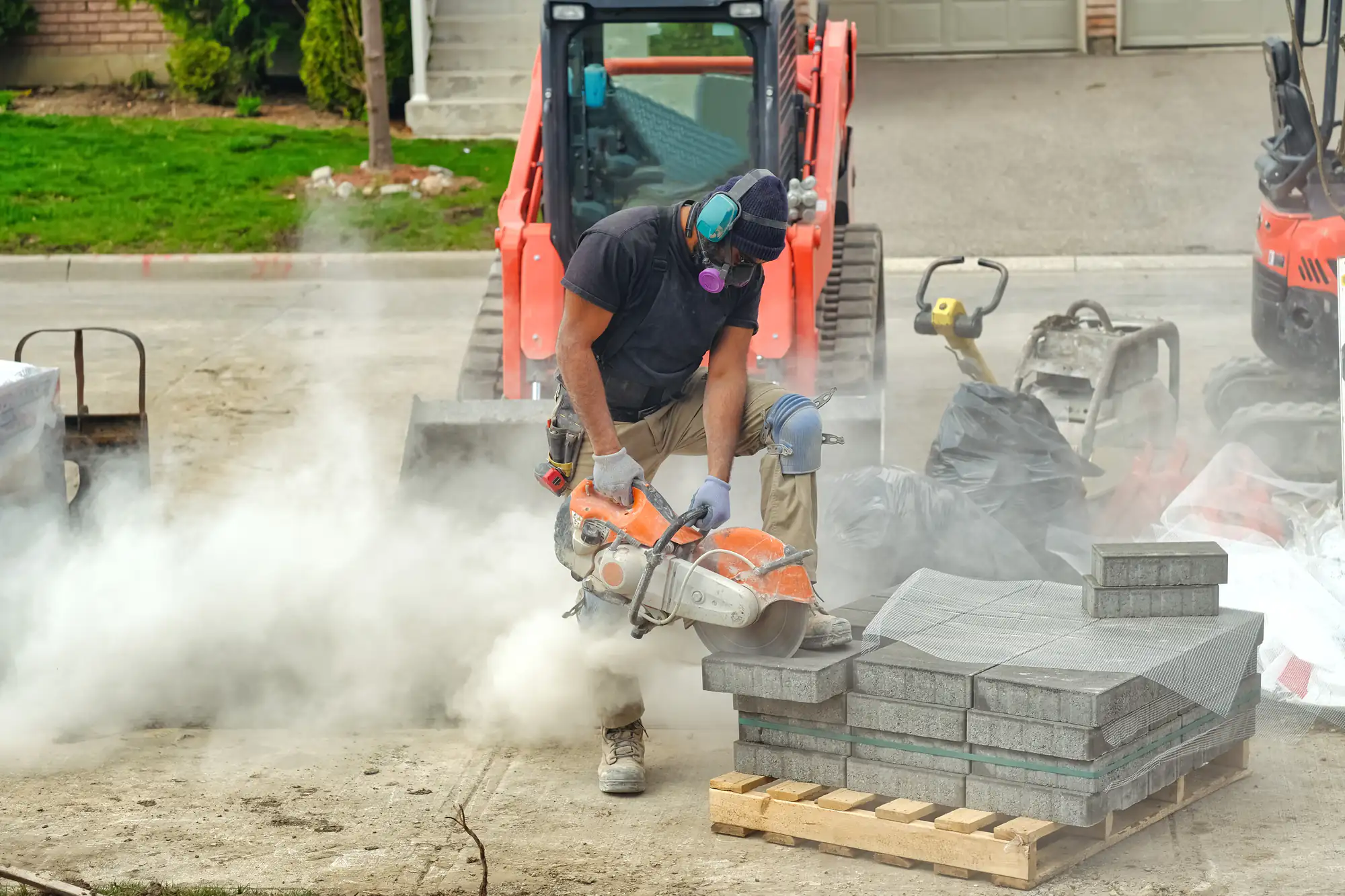 A construction worker wearing a mask and safety goggles uses a power saw to cut through concrete, creating a cloud of dust. Stacks of paving stones and construction equipment are visible around him.