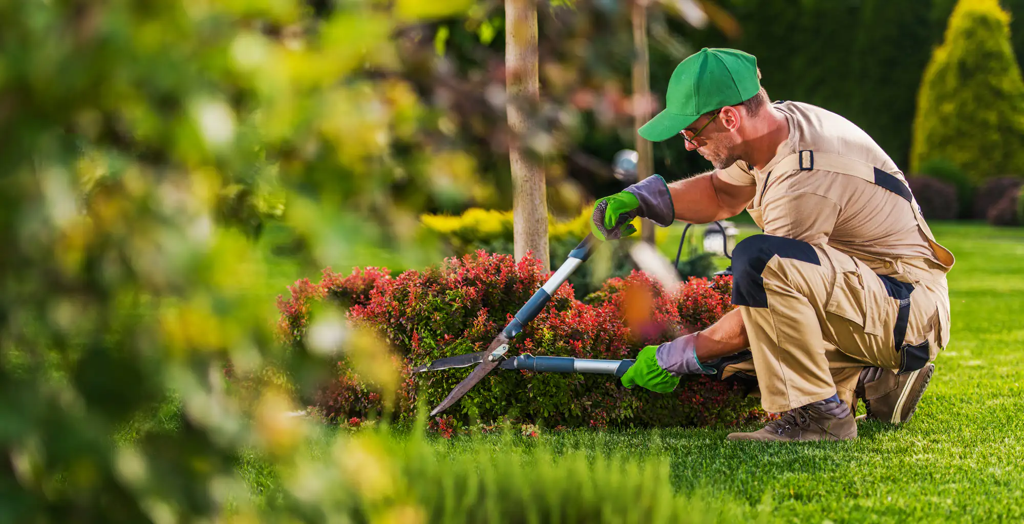 A gardener in khaki overalls, green cap, and gloves kneels on the grass, trimming a bush with hedge shears in a well-maintained garden. Trees and colorful plants are visible in the background.