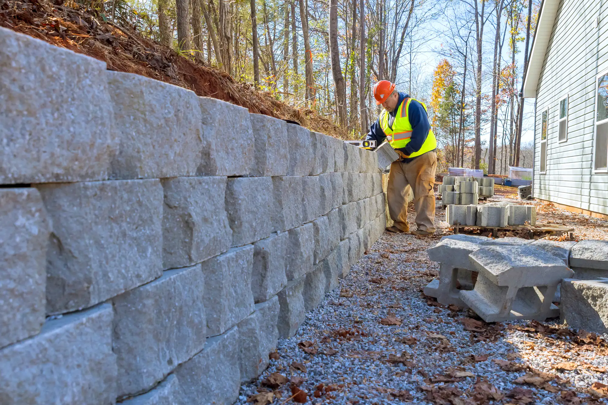 A construction worker in a hard hat and safety vest uses a level to check a stone retaining wall next to a wooded area and house, with pavers and gravel on the ground.